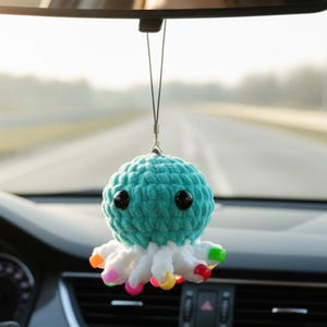 May include: A teal crocheted jellyfish ornament with two large black plastic eyes, hanging from a thin black cord. The base features multiple white tentacles, each tipped with a small, colorful plastic bead in shades of pink, yellow, and green.