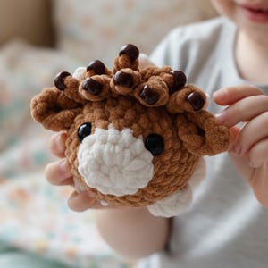 May include: A brown and white crocheted plush toy shaped like a highland cow. The toy features a textured, fuzzy brown body, a white muzzle, two black plastic eyes, and dark brown beads attached to the top of the head to resemble horns.