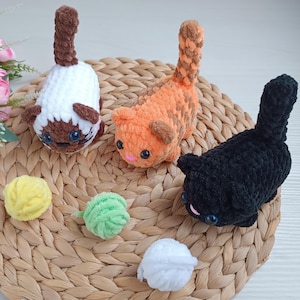 May include: Three crocheted cat toys, one white, one orange, and one black, are displayed on a woven wicker surface. The toys are handmade and have embroidered facial features.