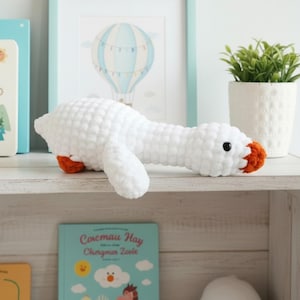 May include: A white crochet goose plush toy with orange feet and beak, and a black eye. The stuffed animal is lying on a white shelf, with a framed picture of a hot air balloon in the background. A small potted plant is also visible.