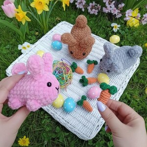 May include: Handmade Easter decorations featuring three plush bunnies in pink, brown, and grey, miniature orange carrots, and decorated eggs. The items are arranged on a white wicker tray, set against a backdrop of green grass and spring flowers.