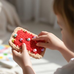 Könnte beinhalten: Ein rotes gehäkeltes Spielzeug, das einer Kuchenscheibe ähnelt, mit einem beigen Kruste und bunten Perlenbelägen. Das Spielzeug wird von einer Person gehalten, deren Finger mit den Perlen interagieren. Das Spielzeug ist für Kinder konzipiert.