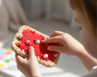 May include: A red crocheted toy resembling a slice of pie, with a tan crust and colorful bead toppings. The toy is held by a person, with their fingers interacting with the beads. The toy is designed for children.