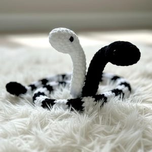 May include: A handmade crochet snake in black and white. The snake has two heads, one white and one black, rising from a body coiled in a circle. The body is black and white checkered. The snake is on a white, fluffy surface.