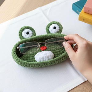 May include: A green crochet frog-shaped glasses holder with white and black eye details, a red mouth, and a white lower lip. The holder contains a pair of eyeglasses. The holder is on a white surface.
