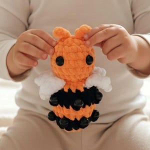 May include: A handmade crochet bee toy. The bee features an orange head and body with black stripes, black bead accents, and white wings. The toy is held by a person.