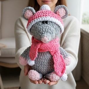 May include: A handmade gray crochet mouse wearing a pink hat and scarf. The hat has a white brim and a white pom-pom. The scarf is pink with white pom-poms at the ends. The mouse has pink ears, nose, and feet. A soft, plush toy.