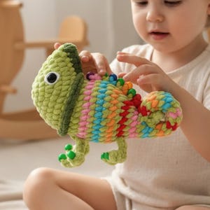 May include: A handmade, colorful crochet chameleon toy. The toy features a green head and feet, with a body of rainbow stripes in shades of pink, blue, orange, and yellow. The chameleon has a large, black and white eye.