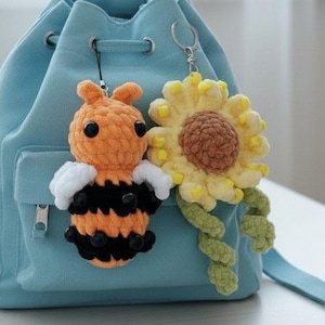 Könnte beinhalten: Ein hellblauer Rucksack mit zwei gehäkelten Schlüsselanhängern. Einer ist eine Biene mit orangefarbenen, schwarzen und weißen Details. Der andere ist eine gelb-braune Sonnenblume mit grünen Blättern. Die Schlüsselanhänger sind am Rucksack befestigt.