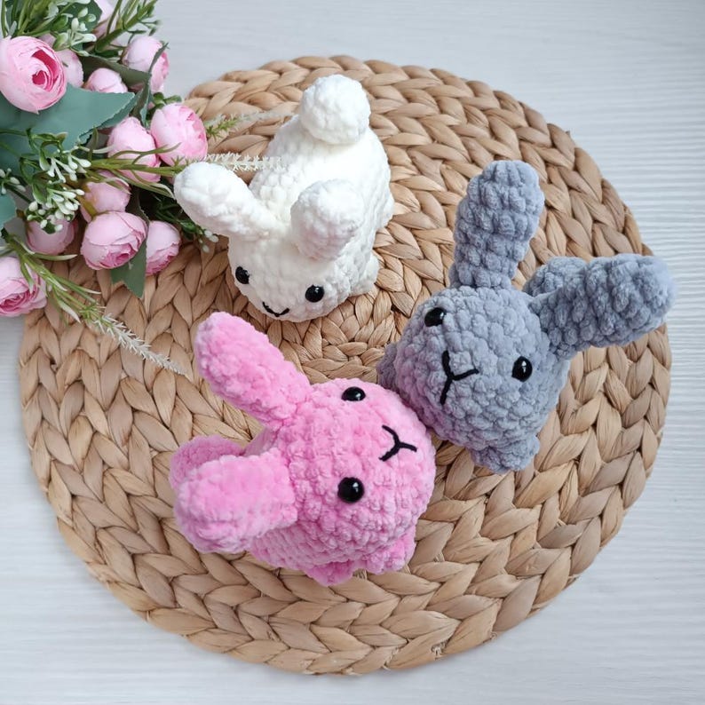 May include: Three handmade crochet bunny rabbits in white, pink, and grey. Each bunny has floppy ears, a small pom-pom tail, and black embroidered facial features. They are arranged on a woven, circular mat, with pink flowers in the background.