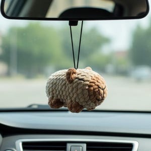 May include: A crocheted capybara car charm in beige and brown, suspended from a rearview mirror. The plush animal has a round body, small ears, and a smiling face. The charm hangs by a black string.