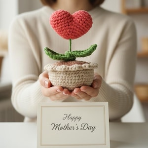 Könnte beinhalten: Eine handgefertigte Häkelblume mit einer roten herzförmigen Blüte, grünen Blättern und einem beigen Topf. Die Blume wird vor einer Karte mit der Aufschrift "Happy Mother's Day" gehalten. Die Häkelblume ist eine einzigartige Geschenkidee.