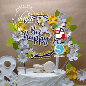 Puede incluir: Un pastel blanco decorado con un adorno de abeja que dice "bee happy". El adorno está rodeado de flores amarillas y blancas y hojas verdes. El pastel también está decorado con perlas blancas y dos macarons.