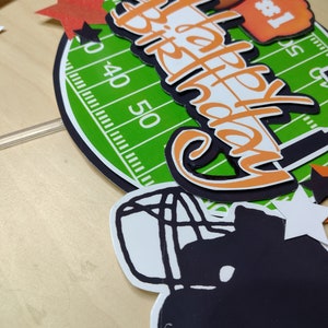 K&ouml;nnte beinhalten: Ein Geburtstagskuchenaufsatz mit Football-Motiv. Das Design umfasst ein gr&uuml;nes Feld, einen Football, einen Schaumstoff-Finger und die Worte "Happy Birthday" in Orange und Wei&szlig;. Dekorative Sterne und ein Helm runden das Design ab.