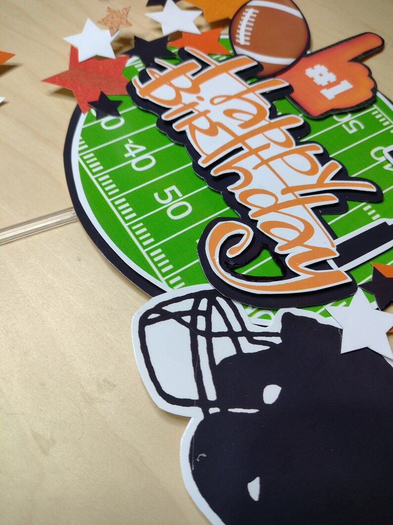 K&ouml;nnte beinhalten: Ein Geburtstagskuchenaufsatz mit Football-Motiv. Das Design umfasst ein gr&uuml;nes Feld, einen Football, einen Schaumstoff-Finger und die Worte "Happy Birthday" in Orange und Wei&szlig;. Dekorative Sterne und ein Helm runden das Design ab.