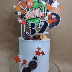 K&ouml;nnte beinhalten: Eine wei&szlig; glasierte Torte mit Football-Dekoration. Die Torte ist mit einem Football, einem Schaumstofffinger und einer Footballfeld-Grafik verziert. Auf der Torte stehen die Worte "Happy Birthday" und die Zahl "32".