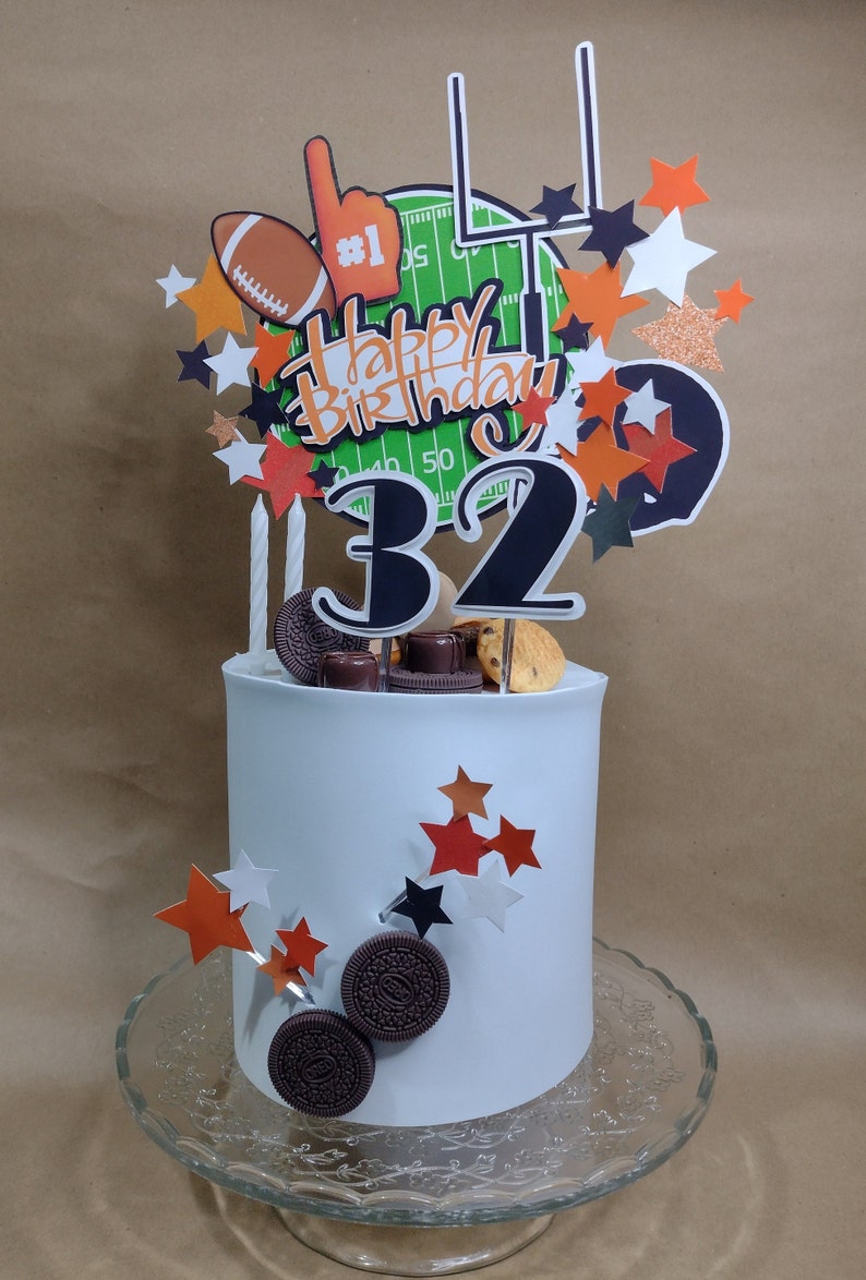 K&ouml;nnte beinhalten: Eine wei&szlig; glasierte Torte mit Football-Dekoration. Die Torte ist mit einem Football, einem Schaumstofffinger und einer Footballfeld-Grafik verziert. Auf der Torte stehen die Worte "Happy Birthday" und die Zahl "32".