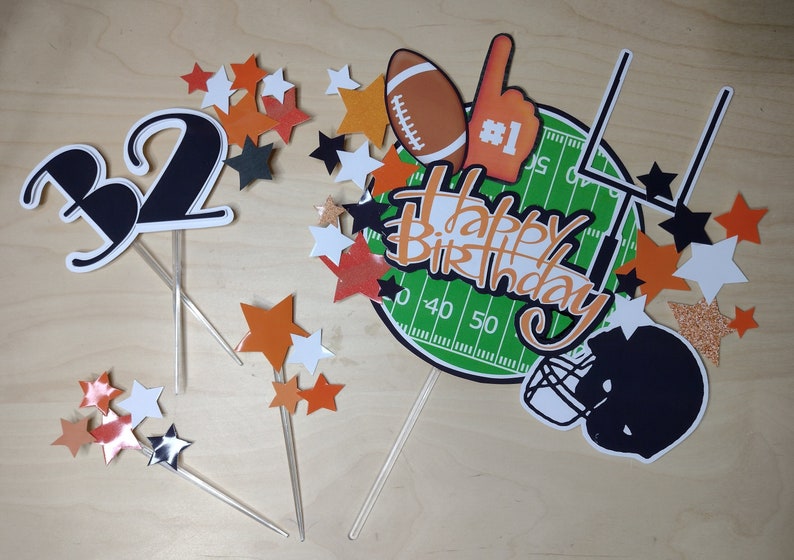 K&ouml;nnte beinhalten: Eine Sammlung von Tortenaufs&auml;tzen zum Thema Football. Enth&auml;lt einen Football, einen Schaumstofffinger, ein Footballfeld, einen Helm und sternf&ouml;rmige Dekorationen. Der Schriftzug "Happy Birthday" ist in Orange und Wei&szlig; gehalten.