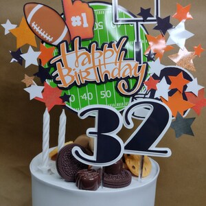 K&ouml;nnte beinhalten: Wei&szlig;er Kuchen mit Football-Dekoration. Der Kuchen ist mit einem Football, einem Schaumstoff-Finger, einem Footballfeld und sternf&ouml;rmigen Dekorationen verziert. Die Aufschrift "Happy Birthday" und die Zahl "32" sind ebenfalls vorhanden.