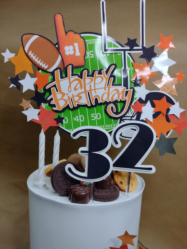 K&ouml;nnte beinhalten: Wei&szlig;er Kuchen mit Football-Dekoration. Der Kuchen ist mit einem Football, einem Schaumstoff-Finger, einem Footballfeld und sternf&ouml;rmigen Dekorationen verziert. Die Aufschrift "Happy Birthday" und die Zahl "32" sind ebenfalls vorhanden.
