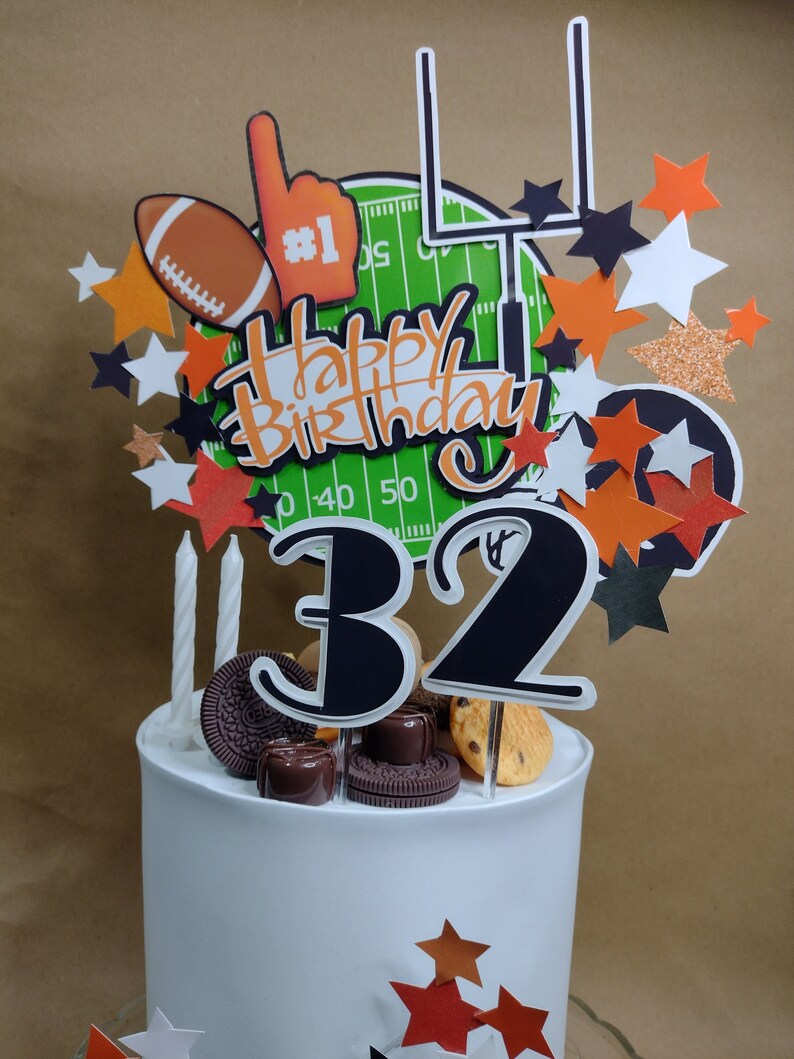 K&ouml;nnte beinhalten: Eine wei&szlig;e Geburtstagstorte mit Fu&szlig;ball-Thema. Die Torte ist mit einem Aufsatz mit einem Football, einem Schaumstofffinger, einem Torpfosten und dem Schriftzug "Happy Birthday" verziert. Die Zahl "32" ist ebenfalls abgebildet.