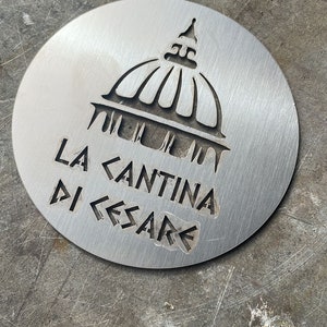 Può includere: Un sottobicchiere in metallo argentato con un design a taglio di un edificio a cupola e il testo "LA CANTINA DI CESARE".