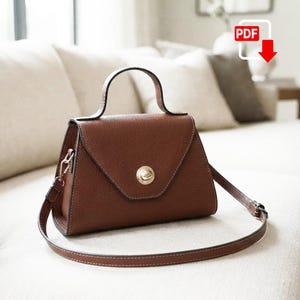 May include: A structured brown handbag with a pebbled texture, featuring a single top handle, a triangular front flap, and a circular gold-tone metal clasp. The bag includes a detachable, adjustable shoulder strap with gold-tone hardware.