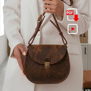 Peut inclure: Un sac besace en cuir marron avec un fond arrondi, un rabat repliable et une fermeture à poussoir en métal doré. Le sac est doté d'une fine bandoulière en cuir réglable avec une boucle dorée et des coutures visibles sur les bords.