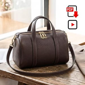Può includere: Una borsa a mano in pelle martellata color marrone scuro a forma di cilindro, dotata di due manici, una tracolla rimovibile e una chiusura a zip in metallo dorato con due tiralampo abbinati. La borsa presenta una struttura con cuciture a vista.
