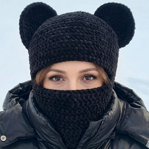 May include: A black knitted hat with two large, round ears and a matching neck warmer. The hat and neck warmer are made of a soft, textured material. The person is wearing a black jacket.
