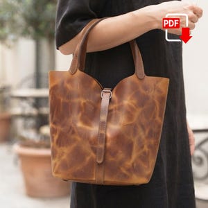 May include: A brown leather bucket bag with a distressed, marbled texture, featuring two top handles and a vertical strap closure with a silver-toned metal buckle. The bag has a structured base and a curved top opening, showcasing a natural finish.