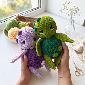 Puede incluir: Dos peluches de tortuga hechos a mano con ganchillo. Uno es verde y verde oscuro, el otro es morado y morado oscuro. Las tortugas tienen ojos negros grandes y extremidades pequeñas. El fondo incluye hilo, flores y una ventana.