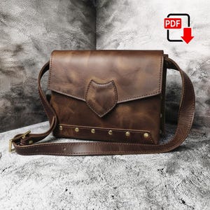 May include: A brown leather shoulder bag featuring a flap closure with a pointed center tab, a row of six brass-colored metal studs along the bottom front, and an adjustable strap with a brass-colored metal buckle. A PDF icon is visible.