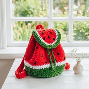 May include: A crochet backpack in the shape of a watermelon, featuring red, green, and white colours. The flap is designed to look like a watermelon slice, with black seed details. The base is green, and the bag has red straps and a drawstring closure.