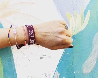 Op de afbeelding: Een set van drie armbanden om de pols: een dunne regenboogkleurige kralenarmband, een lichtroze koordarmband met witte ronde kralen die MASA spellen, en een koraalkleurige kralenarmband naast een brede donkerrode stoffen polsband.