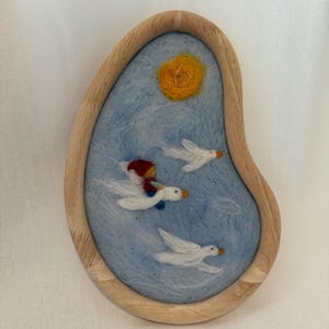 Puede incluir: Una pieza de arte en fieltro caprichosa que representa un grupo de gansos blancos volando sobre un cielo azul con un sol amarillo. Un ganso lleva una pequeña figura. La obra está enmarcada en madera.
