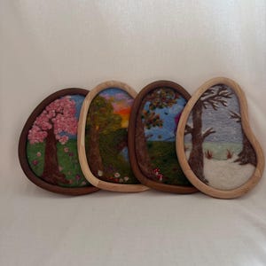 Op de afbeelding: Vier vilten landschapskunstwerken met houten frames. Elk stuk toont een ander seizoen: lente met roze bloesem, zomer met een zonsondergang, herfst met rode paddenstoelen en winter met sneeuw. De frames zijn ovaal.