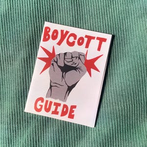Puede incluir: Un folleto blanco con las palabras "BOYCOTT GUIDE" en rojo, con un puño gris y estrellas rojas. El folleto está sobre una superficie de pana verde.