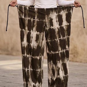 May include: Wide-leg pants with a brown and white tie-dye pattern. The pants are held up by a person wearing a white long-sleeve shirt. The pants have a drawstring waist and are made of a soft, flowing fabric.