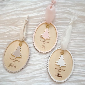 K&ouml;nnte beinhalten: Drei ovale Weihnachtsanh&auml;nger aus Holz mit gewelltem Rand, jeweils mit einem kleinen goldenen Glitzerstern, einem ausgeschnittenen Baum und dem Text "Mon Premier No&euml;l 2025" sowie den Namen Giulian, Iris oder Alma, mit Stoffb&auml;ndern.