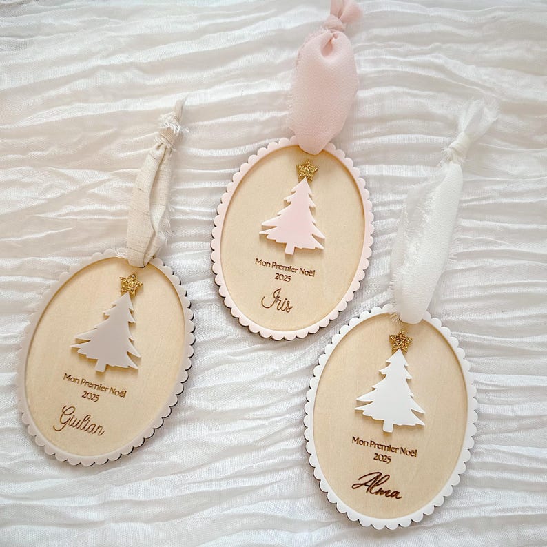 K&ouml;nnte beinhalten: Drei ovale Weihnachtsanh&auml;nger aus Holz mit gewelltem Rand, jeweils mit einem kleinen goldenen Glitzerstern, einem ausgeschnittenen Baum und dem Text "Mon Premier No&euml;l 2025" sowie den Namen Giulian, Iris oder Alma, mit Stoffb&auml;ndern.
