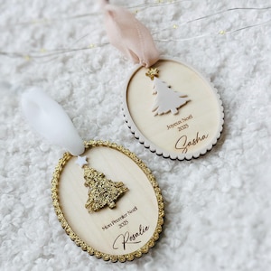 K&ouml;nnte beinhalten: Zwei ovale Weihnachtsanh&auml;nger aus Holz mit gewelltem Rand. Einer zeigt einen goldglitzernden Baum, ein wei&szlig;es Band und den Text Mon Premier Noel 2025 Rosalie. Der andere hat einen wei&szlig;en Baum, ein rosafarbenes Band und den Text Joyeux Noel 2025 Sasha.