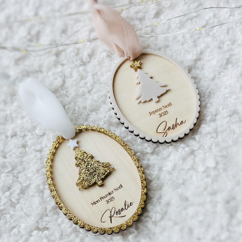 K&ouml;nnte beinhalten: Zwei ovale Weihnachtsanh&auml;nger aus Holz mit gewelltem Rand. Einer zeigt einen goldglitzernden Baum, ein wei&szlig;es Band und den Text Mon Premier Noel 2025 Rosalie. Der andere hat einen wei&szlig;en Baum, ein rosafarbenes Band und den Text Joyeux Noel 2025 Sasha.