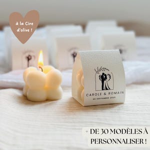 Può includere: Scatole di bomboniere bianche con una piccola candela bianca all'interno. Le scatole sono decorate con una silhouette nera di una coppia e il testo "CAROLE & ROMAIN 20 SEPTEMBRE 2025". Il testo "à la Cire d'olive!" è scritto a forma di cuore sopra le scatole.