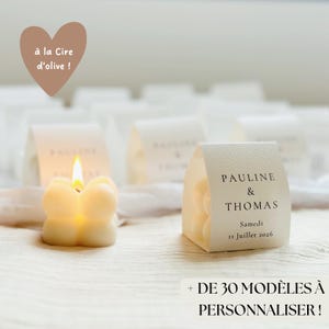 Könnte beinhalten: Eine brennende, elfenbeinfarbene Kerze mit einer einzigartigen, abgerundeten Form, neben einer kleinen, gefalteten Papierbox. Die Box zeigt die Namen "PAULINE & THOMAS" und das Datum "11 Juillet 2026". Der Text "+ DE 30 MODÈLES À PERSONNALISER!" ist ebenfalls enthalten.