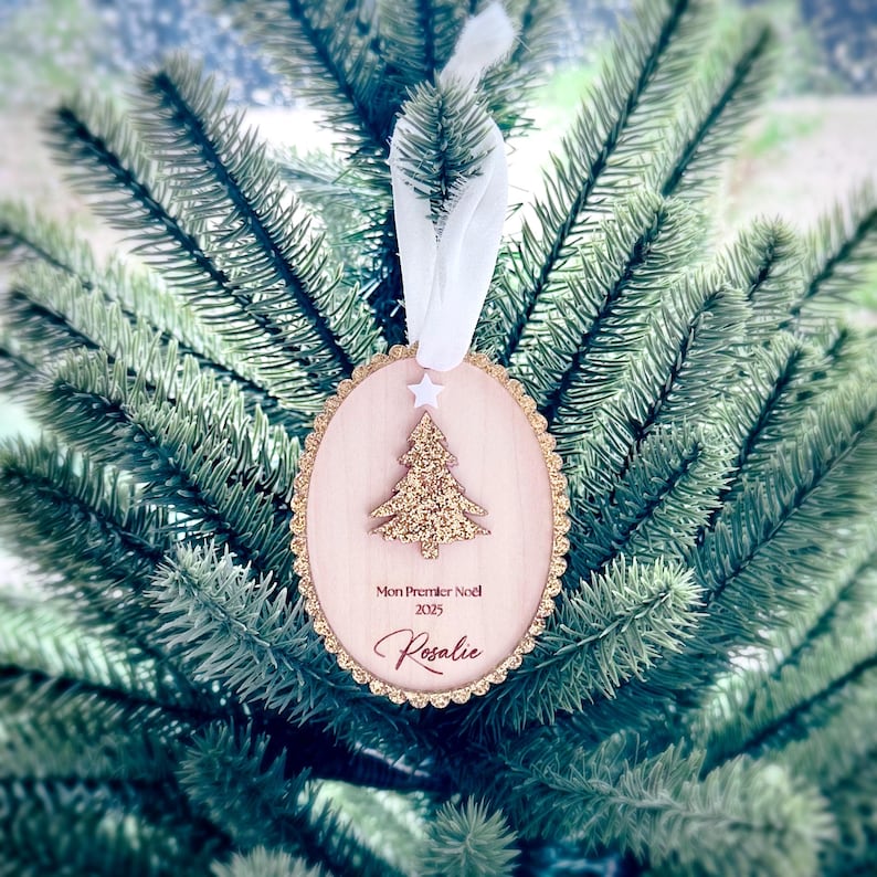 K&ouml;nnte beinhalten: Ein ovaler Weihnachtsbaumanh&auml;nger aus Holz mit einem gewellten Rand aus Goldglitter, verziert mit einem goldglitzernden Weihnachtsbaum, einem kleinen wei&szlig;en Stern und dem Text Mon Premier No&euml;l 2025 Rosalie, an einem wei&szlig;en Band.