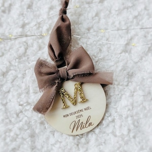 Peut inclure: Ornement de Noël en bois avec un ruban et un nœud marron. L'ornement présente la lettre "M" en paillettes dorées et le nom "Mila" en cursive, ainsi que le texte "MON DEUXIÈME NOËL 2025".
