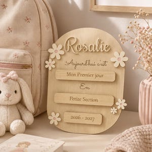 Puede incluir: Un letrero ovalado de madera con el nombre Rosalie en relieve, junto al texto en francés Aujourd'hui c'est, Mon Premier jour, En, Petite Section y 2026-2027. El letrero está decorado con pequeñas flores de madera de color claro.