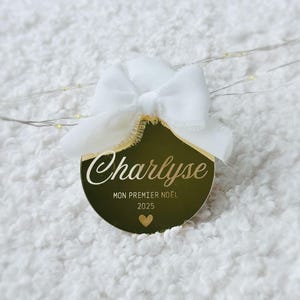Puede incluir: Un adorno navideño dorado y redondo con el nombre "Charlyse" en escritura cursiva. El adorno también presenta el texto "MON PREMIER NOËL 2025" y un pequeño corazón. Una cinta blanca está atada en la parte superior.
