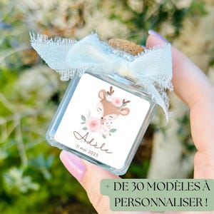 Può includere: Una piccola bottiglia di vetro con un tappo di sughero e un nastro bianco legato intorno. La bottiglia ha un'etichetta con un cervo dei cartoni animati con fiori rosa e il nome "Adèle" e la data "11 mai 2025". Il testo "DE 30 MODÈLES À PERSONNALISER!" è visibile nell'angolo in basso a destra dell'immagine.